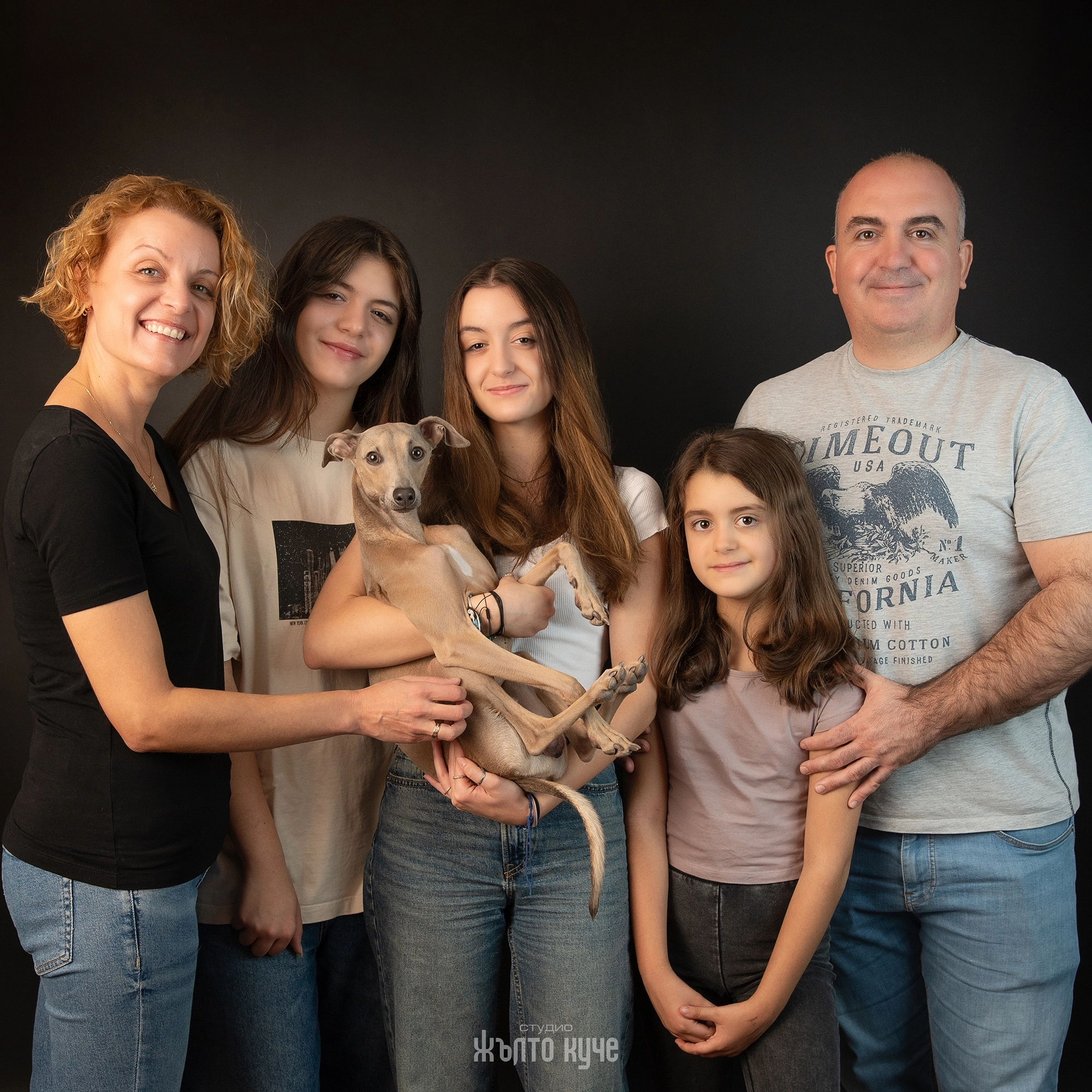 Щастливо семейство от петима души позира за студийна фотосесия с италиански грейхаунд, държан в прегръдките на едно от момичетата, на черен фон.