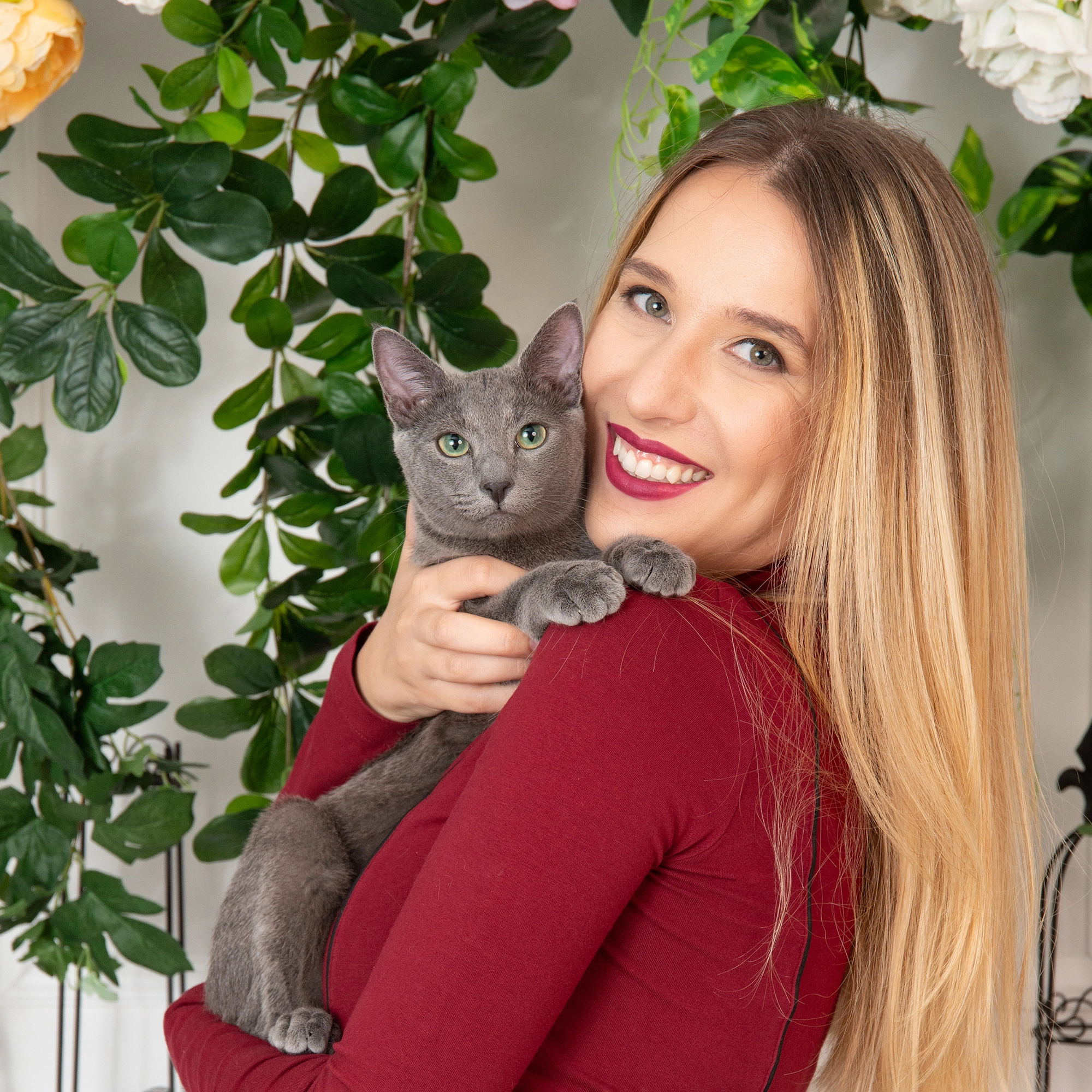 Млада жена с дълга руса коса и усмивка, държаща сива котка в прегръдките си, на фона на зелени растения и цветя.